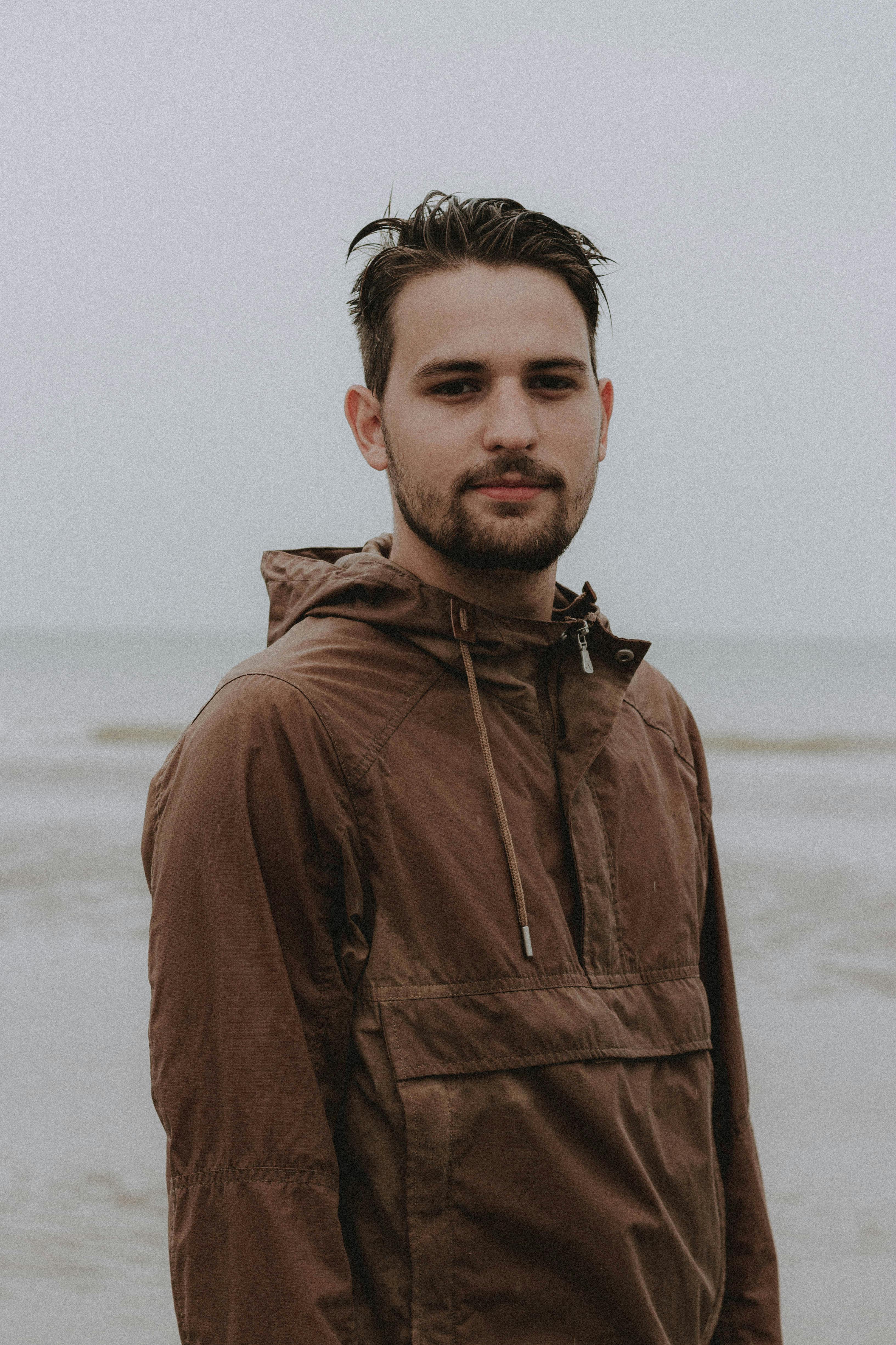Bearded male with wet hair in brown jacket standing near waving sea in overcast weather while looking at camera on seaside