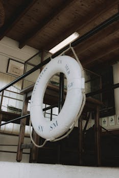 Low angle of white lifebuoy with inscription hanging on metal railing on ropes under wooden ceiling in harbor in daylight