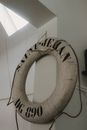 Shabby white lifebuoy with cords hanging on metal chain against white wall near window in daytime in light room inside