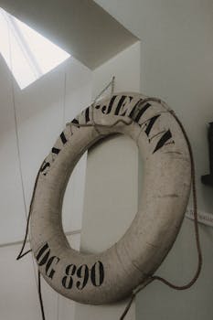 Shabby white lifebuoy with cords hanging on metal chain against white wall near window in daytime in light room inside