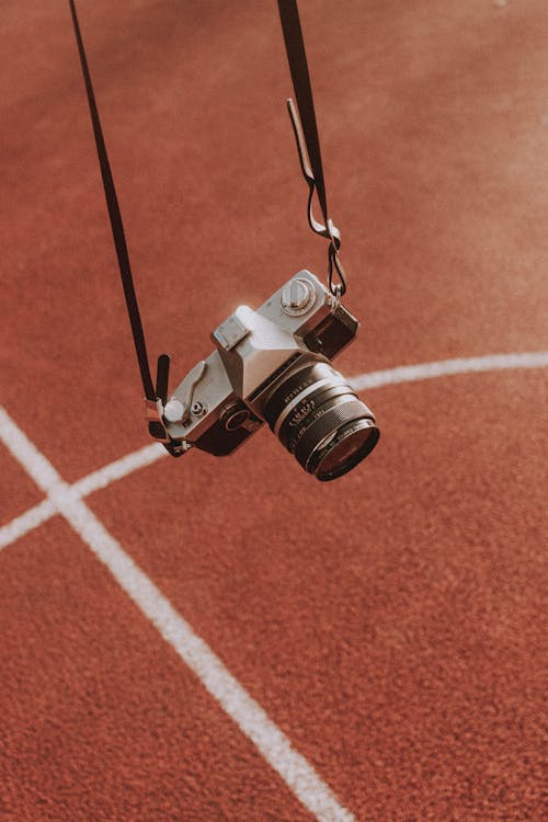 Old fashioned photo camera hanging on black strap against red ground with marking on stadium in daylight in summer day