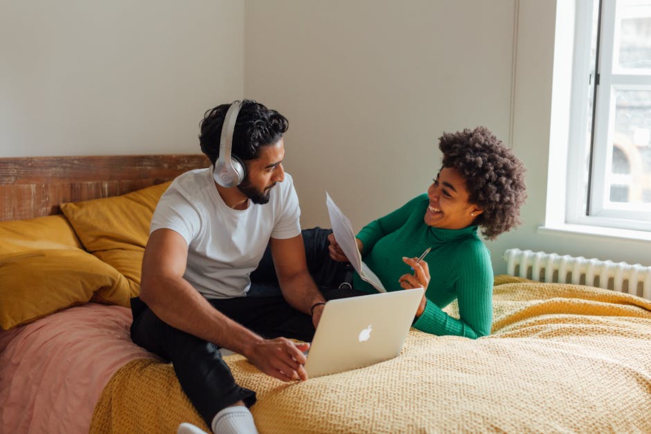 A joyful interracial couple enjoying leisure time with a laptop in a cozy bedroom setting.