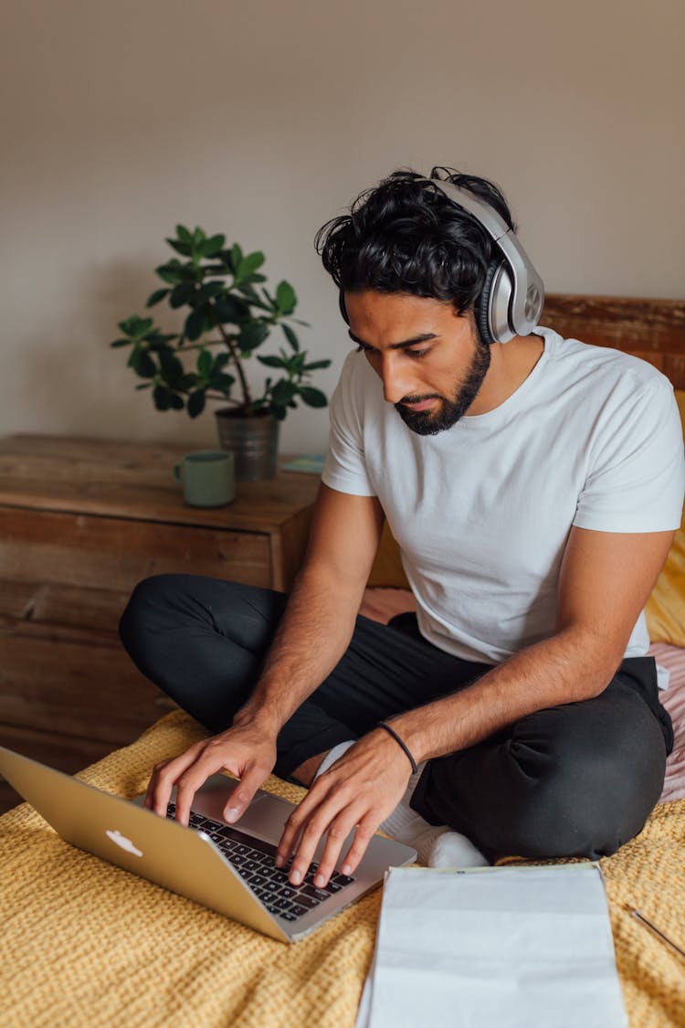 A Man In White Shirt Sitting On The Bed While Working On His Laptop