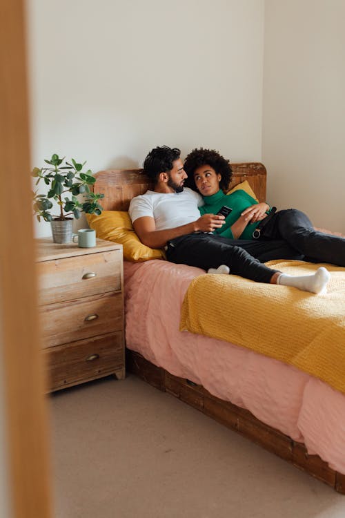 Free Woman in Green Long Sleeve Shirt and Black Pants Lying on Bed Beside a Man Stock Photo