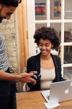 A smiling woman uses a pay terminal at her work desk with a laptop in a cozy indoor setting.
