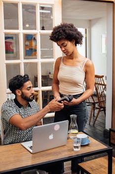 Man at laptop discusses payment with woman using a card reader in a cozy cafe.