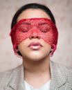 Enigmatic female with pink lip gloss and black hair standing in light room near wall with red mesh textile on closed eyes
