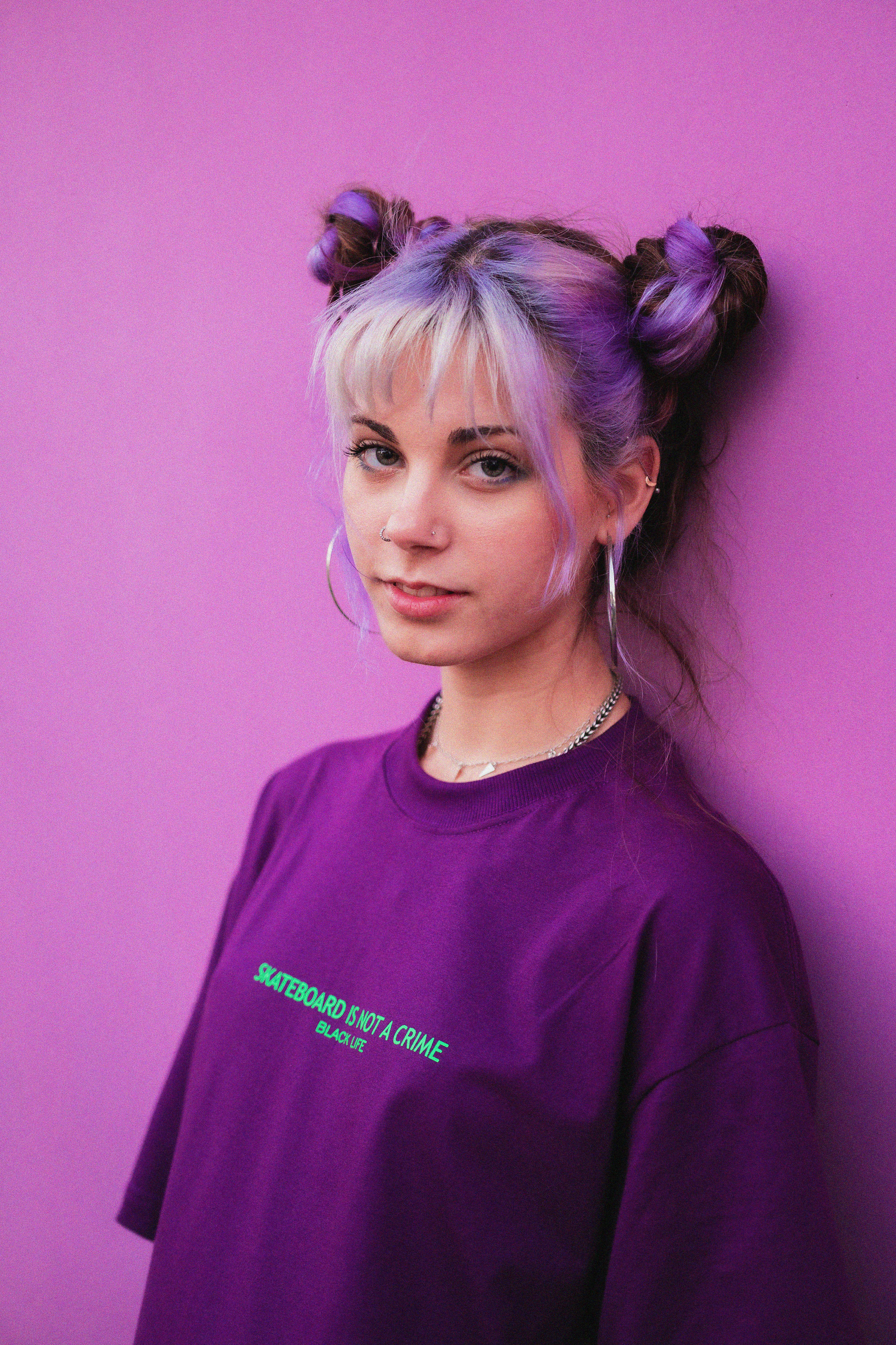 Female in trendy purple t shirt and with dyed hairstyle leaning on bright wall and looking at camera on purple background