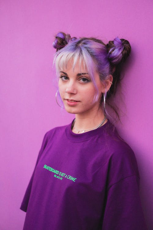 Female in trendy purple t shirt and with dyed hairstyle leaning on bright wall and looking at camera on purple background