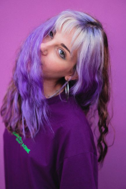 Young female with wavy dyed hairstyle and earrings in casual teenage sweater looking at camera on purple background