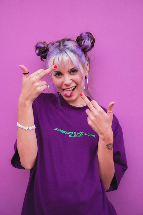 Naughty female teenager demonstrating middle fingers with tongue out and looking at camera on purple background