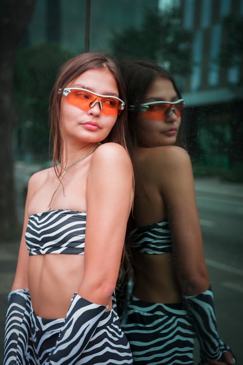 Confident young female in stylish sunglasses and clothes revealing body leaning on reflecting wall