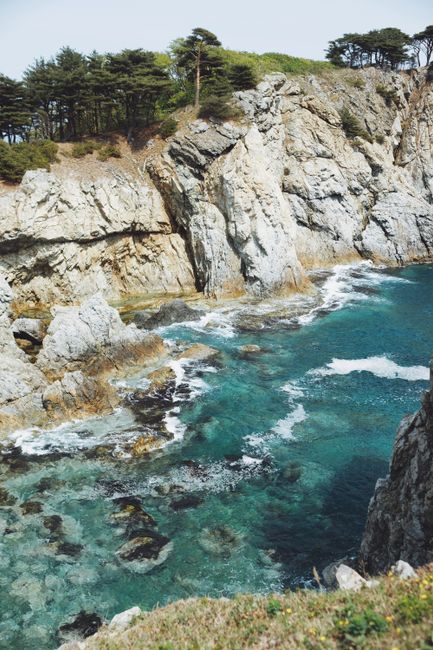 Panoramic view of rocky cliffs meeting turquoise sea waves with lush vegetation.