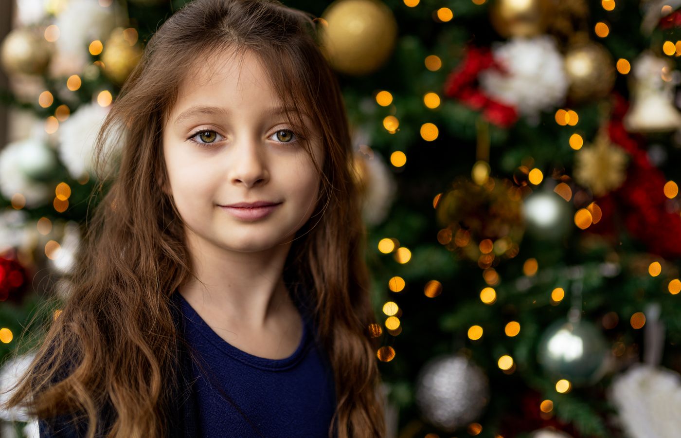 A young girl with wavy brown hair smiles warmly in front of a decorated Christmas tree.