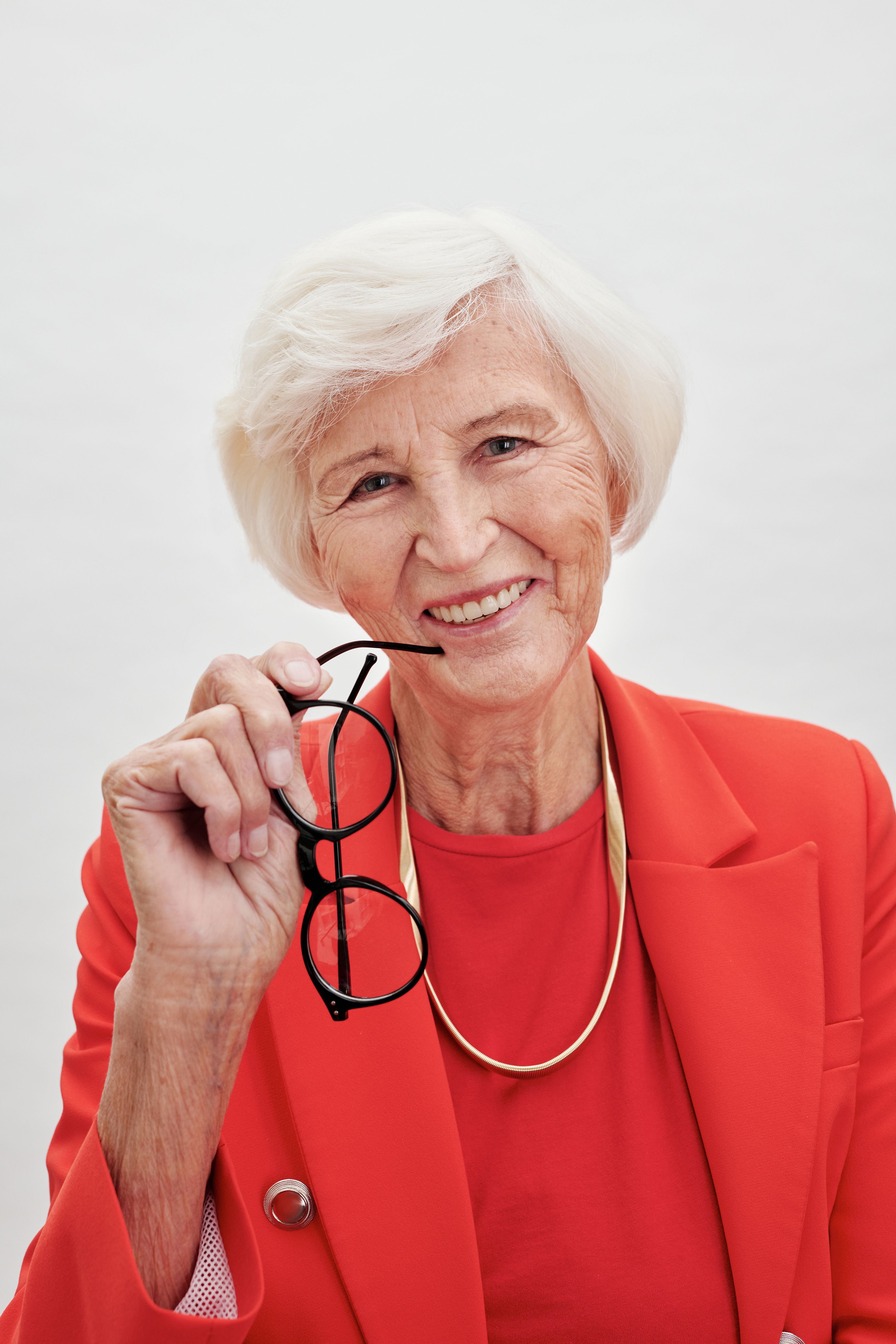 Smiling senior woman in coral red blazer holding glasses, showcasing elegance and style.