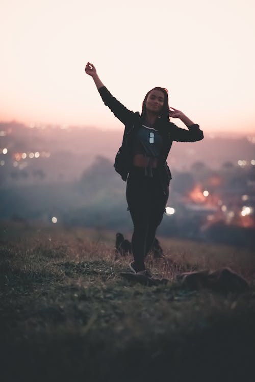 Full body of young slender African American female in casual clothing touching hair and raising hand while walking on lawn near town at sunset on blurred background