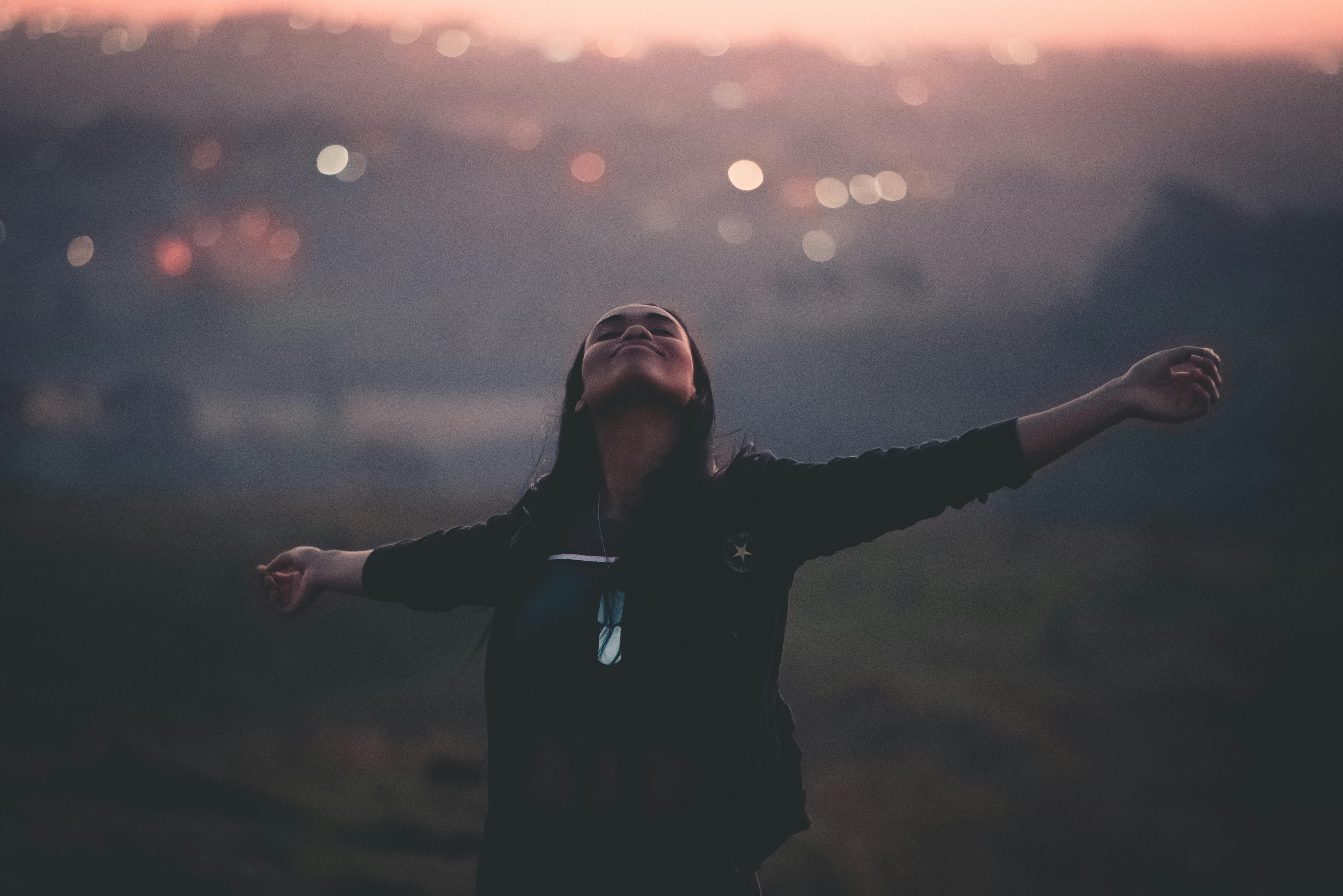Cheerful young African American female outstretching arms with eyes closed while walking in field next to settlement on blurred background
