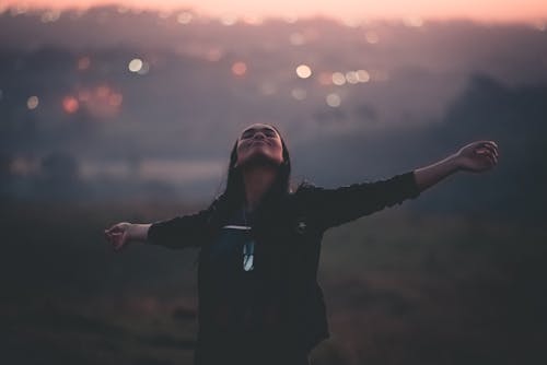 Cheerful young African American female outstretching arms with eyes closed while walking in field next to settlement on blurred background