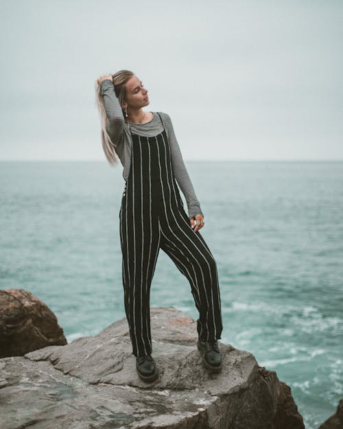 Full body of young confident female traveler in stylish outfit touching long hair while relaxing on rocky boulder and admiring wavy ocean