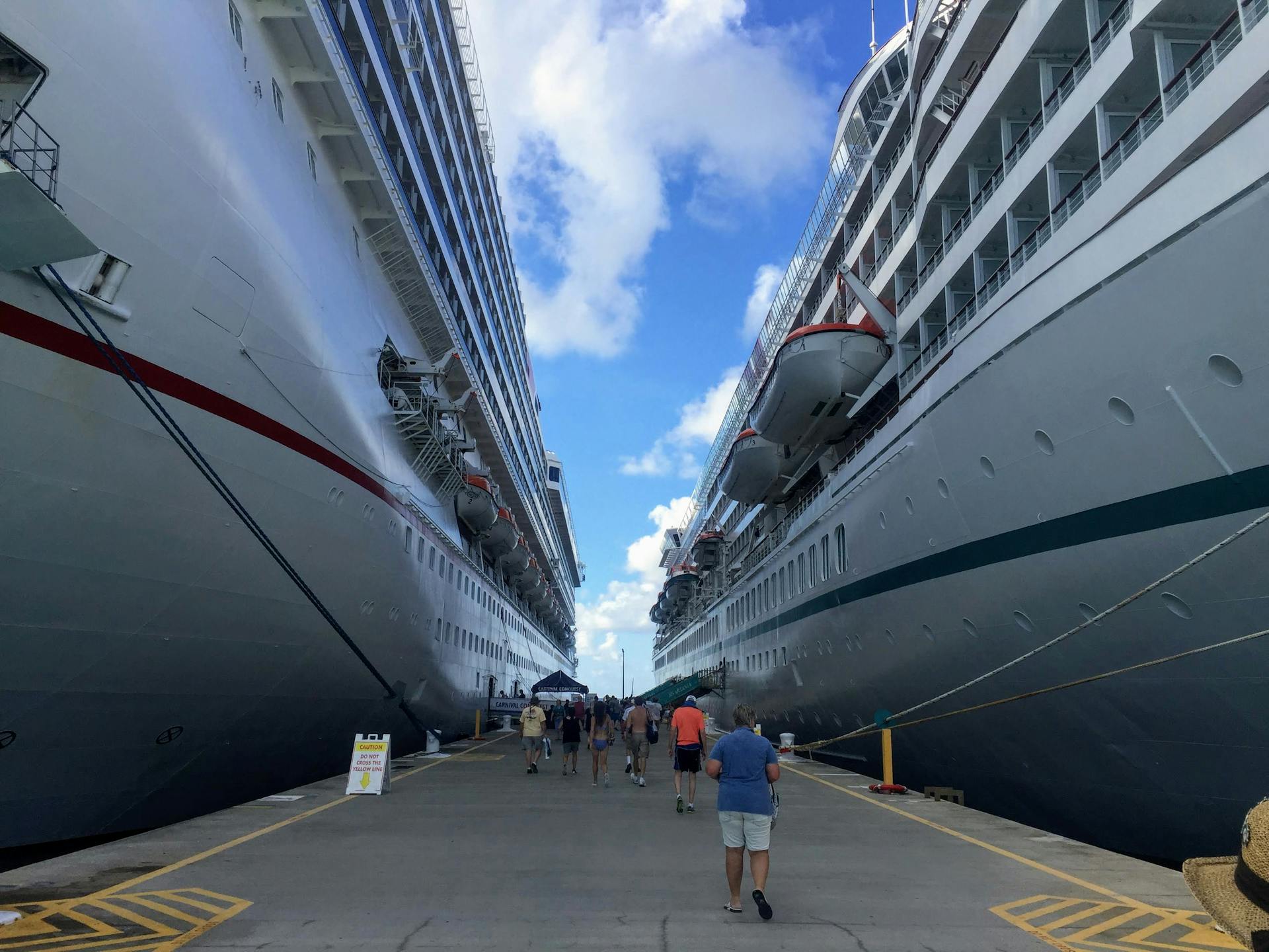 Tourists walking between massive cruise ships in a sunny harbor, enjoying a vacation experience.