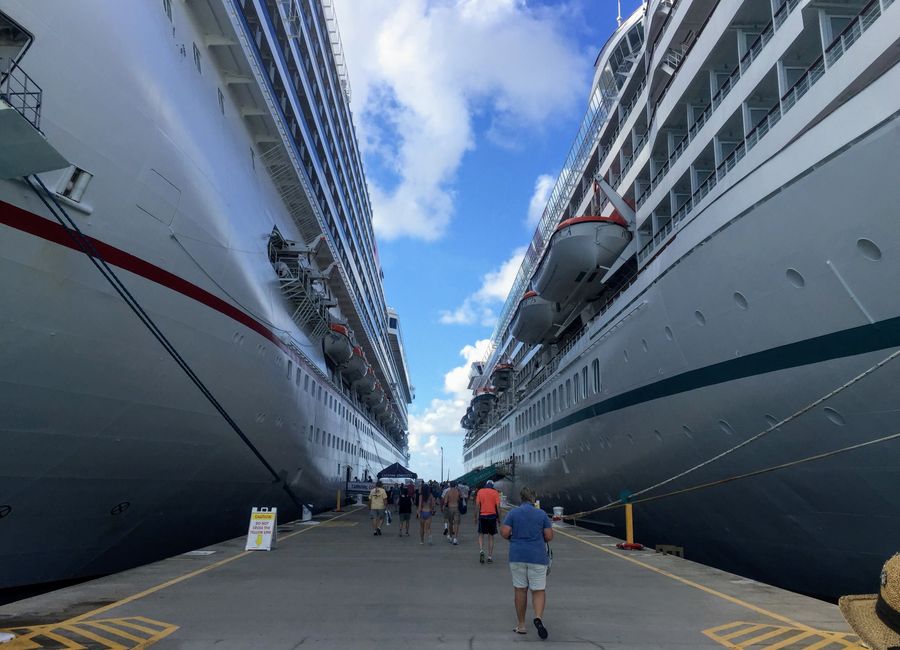 Tourists walking between massive cruise ships in a sunny harbor, enjoying a vacation experience.
