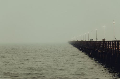 Long pier with streetlights running away above calm sea water in mist under cloudy gloomy sky