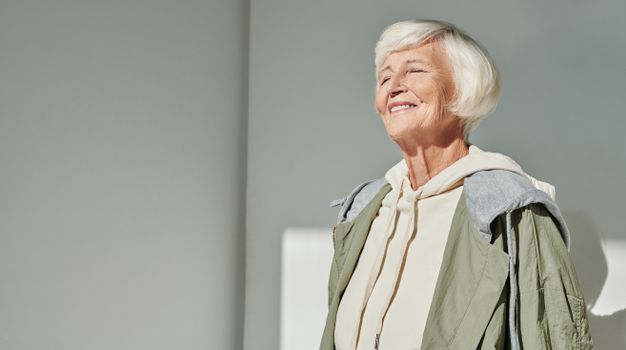 Elegant senior woman smiling in sunlight, wearing stylish casual attire indoors.