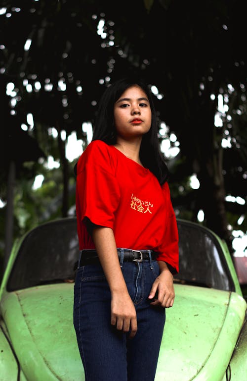 Woman in Red Crew Neck T-shirt and Blue Denim Jeans Standing Beside Green Vintage Car
