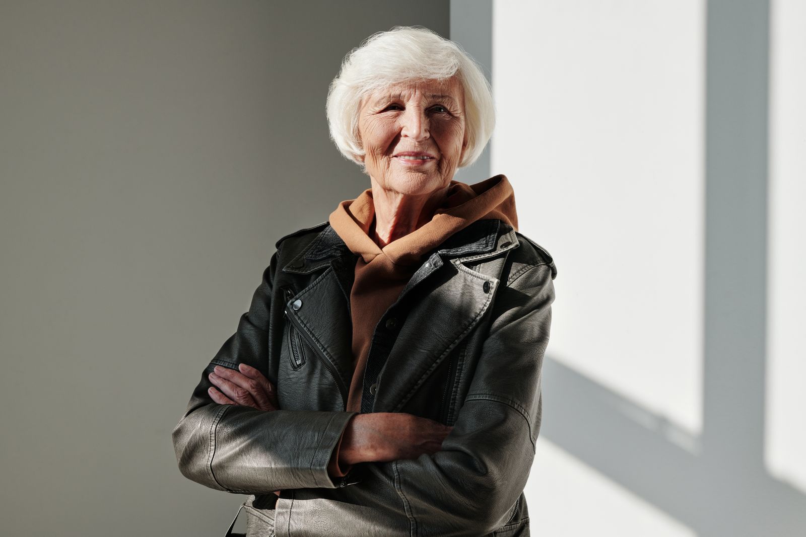 Smiling older woman with white hair, arms crossed, wearing a black leather jacket in warm window light