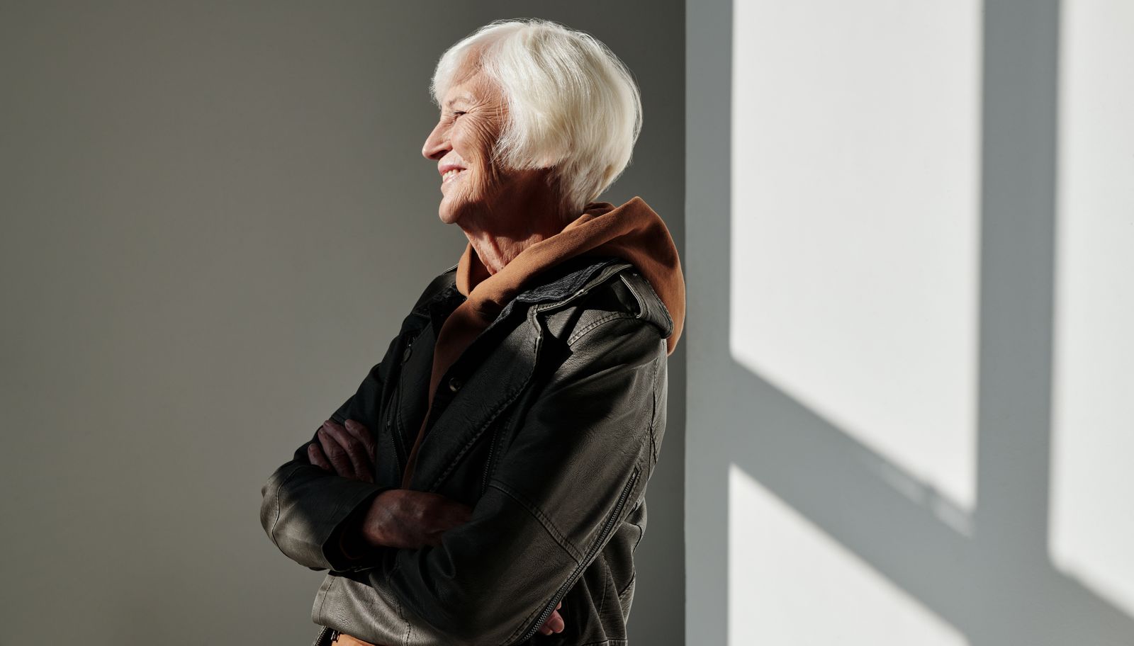 Smiling older woman with white hair and arms crossed stands in warm sunlight beside a pale wall