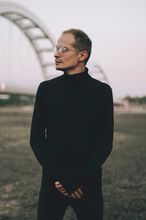 Young dreamy male in black outfit and eyeglasses standing on grass meadow while looking away