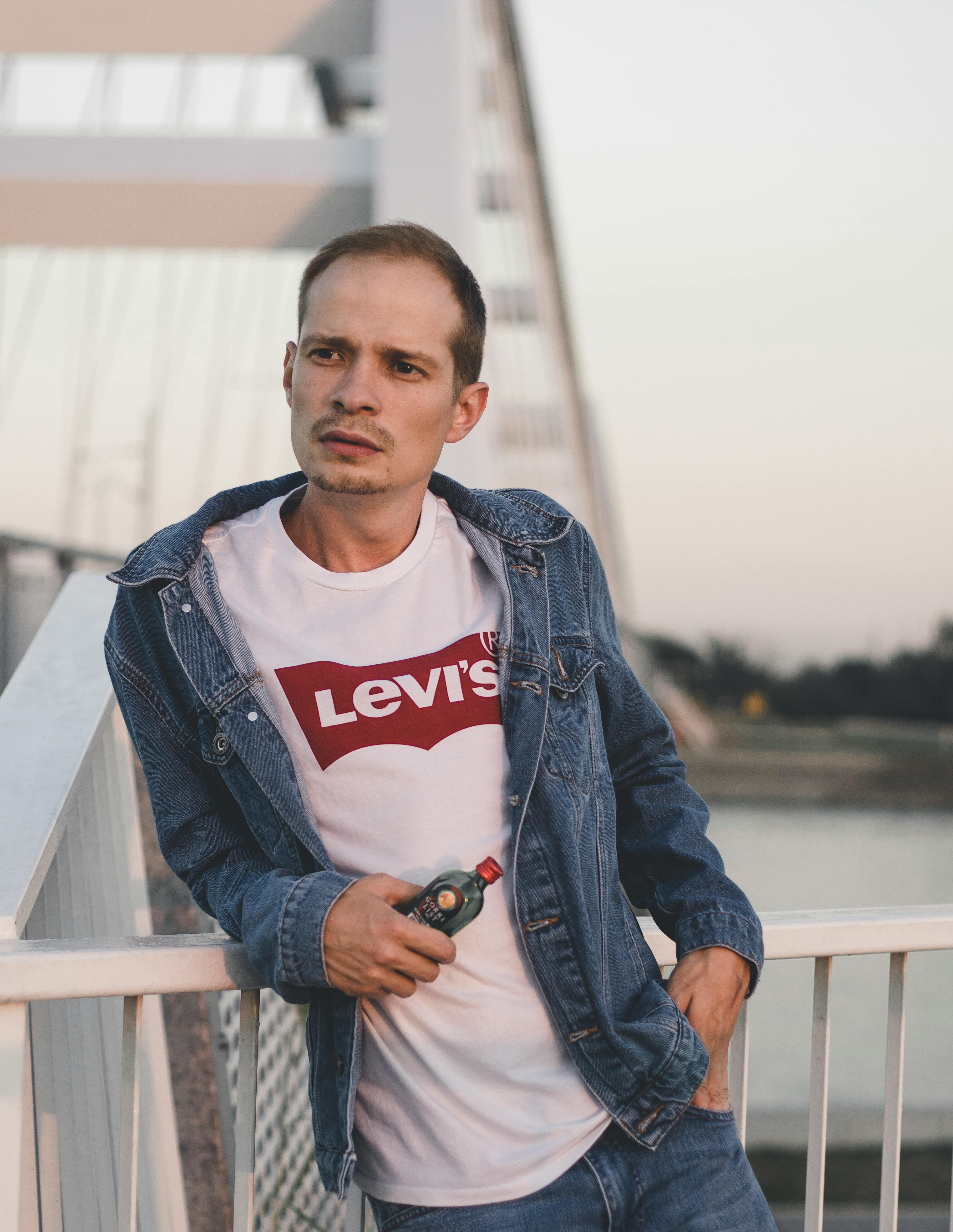Young contemplative male in trendy apparel with bottle of drink looking away while standing with hand in pocket near fence