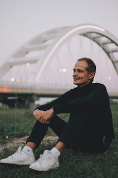 一位穿着时尚黑色服装和运动鞋的年轻男性，快乐地坐在城市桥附近的草地上，目光移向别处
