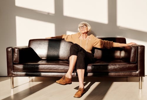 Elderly Woman in Brown Long Sleeve Shirt and Black Pants Sitting on Black Leather Couch