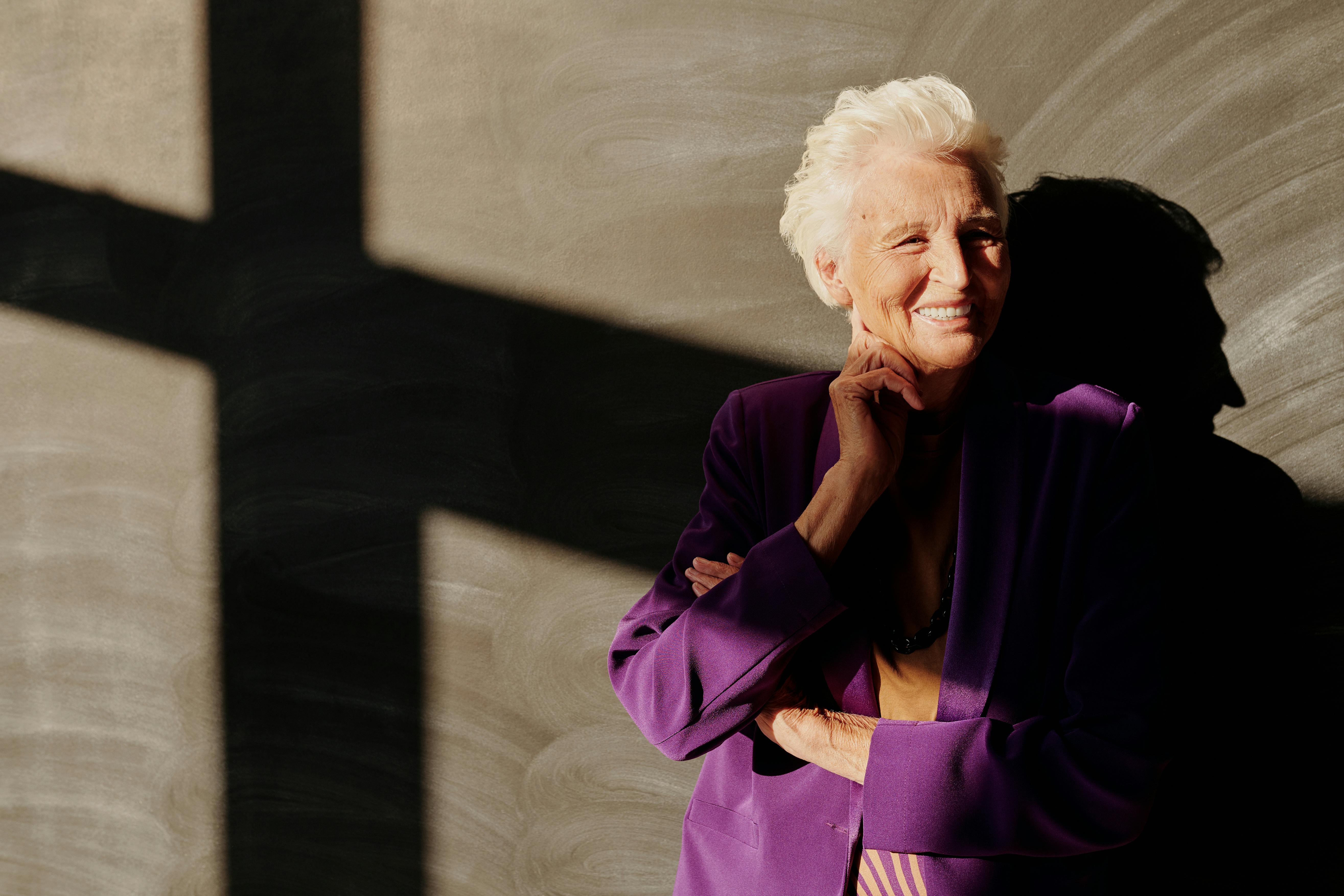 Portrait of an elderly woman in purple, smiling warmly with crossed arms in sunlight.