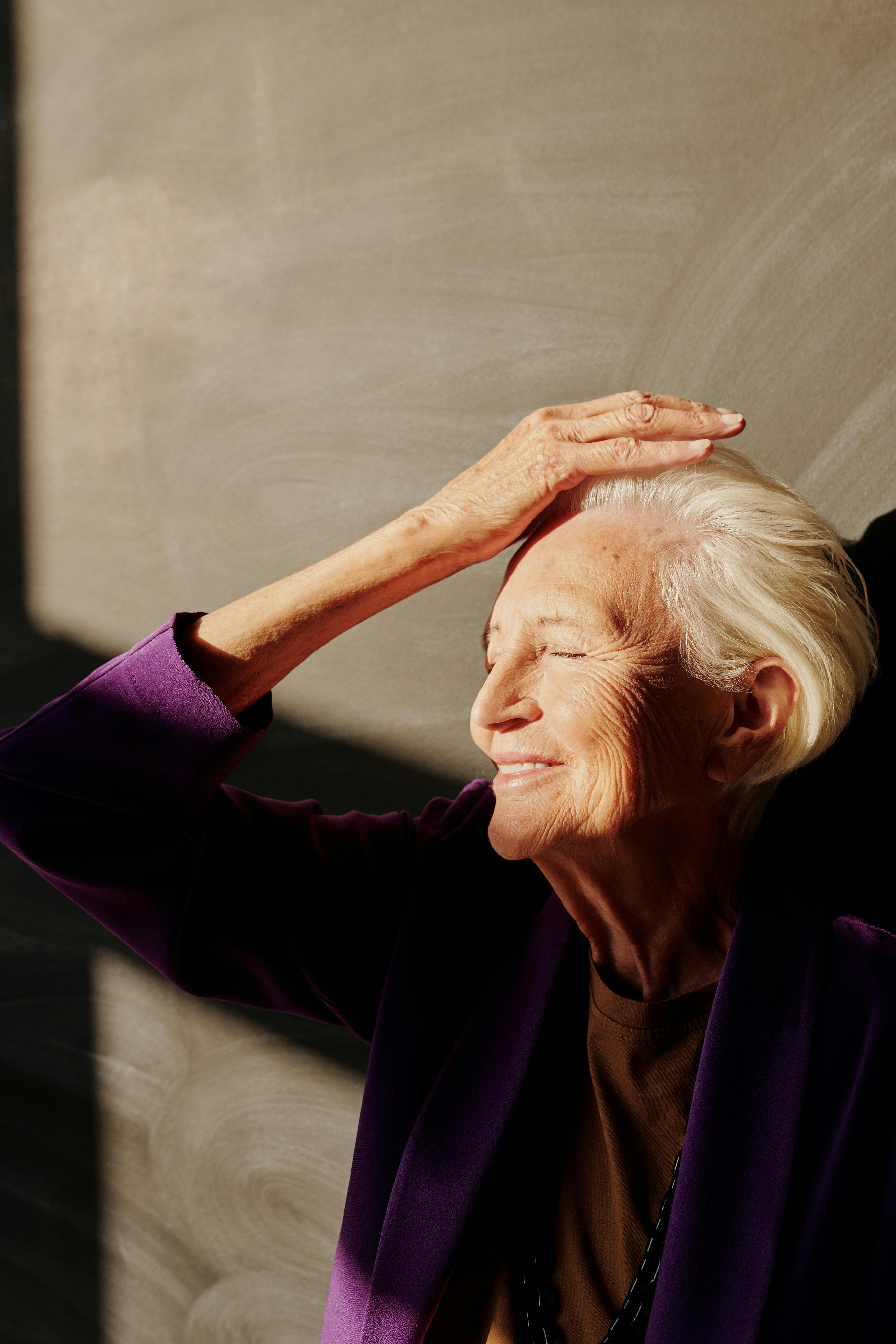 Elderly woman with gray hair smiling, enjoying sunlight indoors.