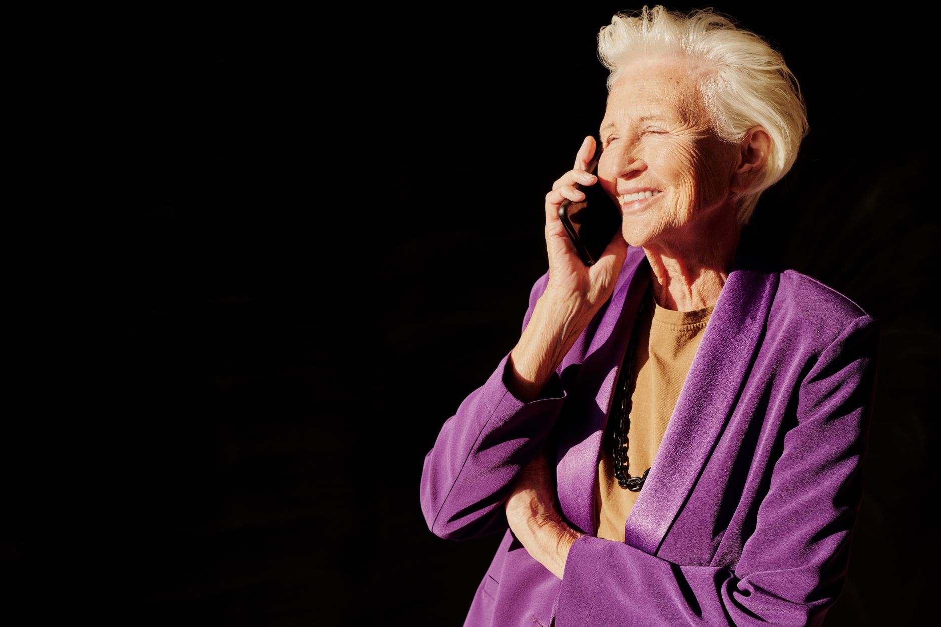 Senior woman in a purple jacket smiling while talking on the phone against a dark background.