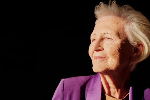 Elegant senior woman smiling with confidence in a purple jacket against black background.