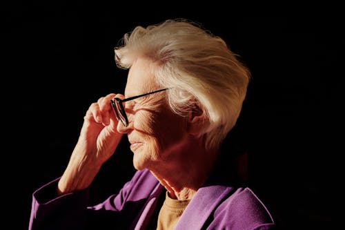 Side profile of a senior woman in a purple jacket, adjusting her glasses against a black background.