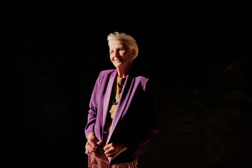 Portrait of a senior woman confidently smiling in a purple blazer against a dark background.