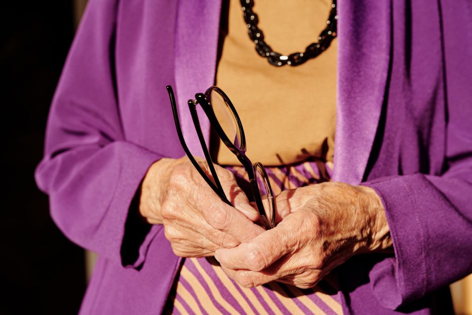 An elderly woman in purple clothing holds eyeglasses, highlighting age and elegance.