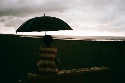 Back view of anonymous female with umbrella enjoying sea on sandy shore in cloudy day