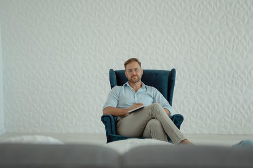 Free A thoughtful man sitting cross-legged in an armchair against a stylish white background. Stock Photo