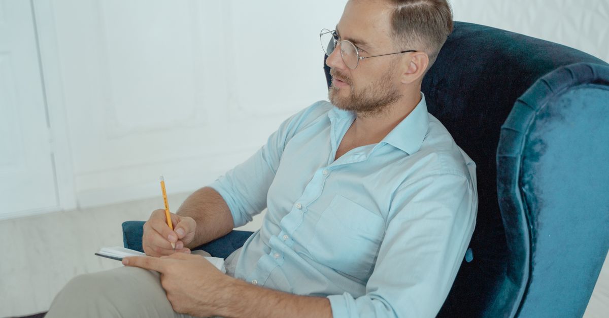 A therapist in a session, wearing glasses and taking notes while seated on an armchair.