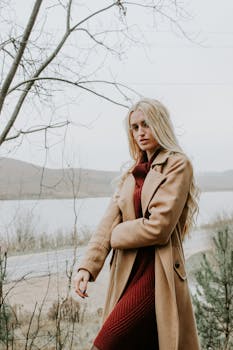 Stylish blonde woman wearing a coat by a lake in autumn, showcasing fashion and nature.