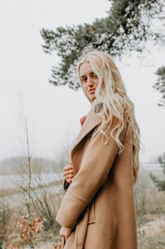 A fashionable blonde woman poses in a winter coat amidst a serene outdoor setting.
