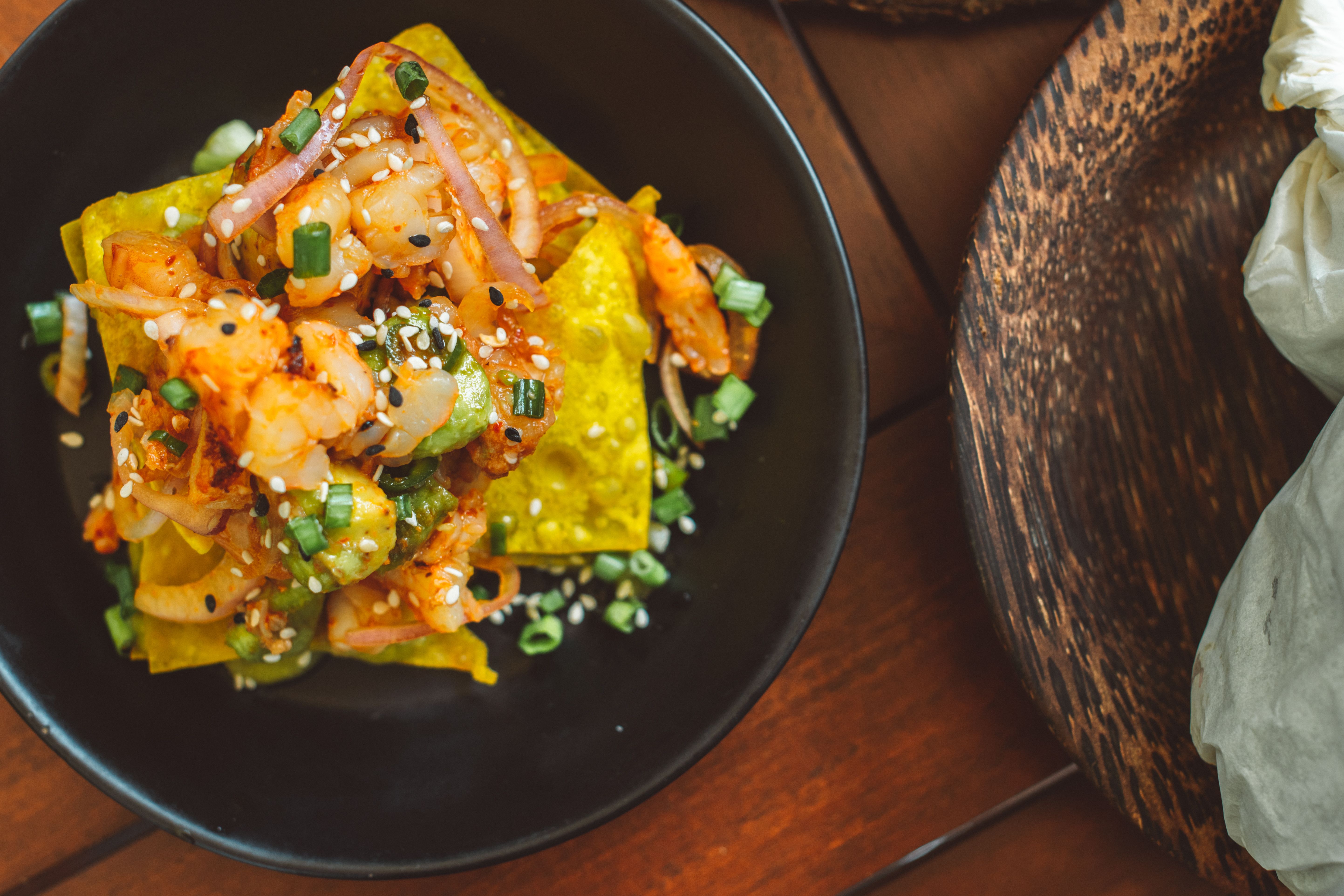 Mouthwatering shrimp and avocado nachos with sesame seeds, onions, and garnishes in a black bowl.