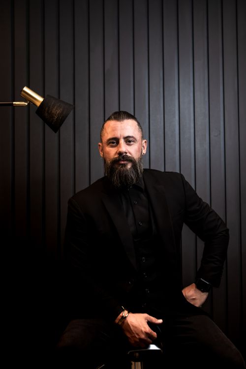 Free Portrait of a bearded man in a black suit, sitting in a modern setting with minimalist decor. Stock Photo