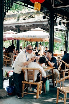 A man performs traditional ear cleaning at an outdoor tea house with bamboo chairs and tables.