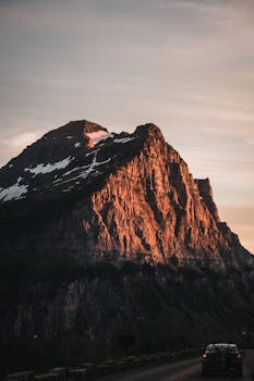 Car on a road with a majestic rocky mountain peak at dusk, reflecting sunset light.